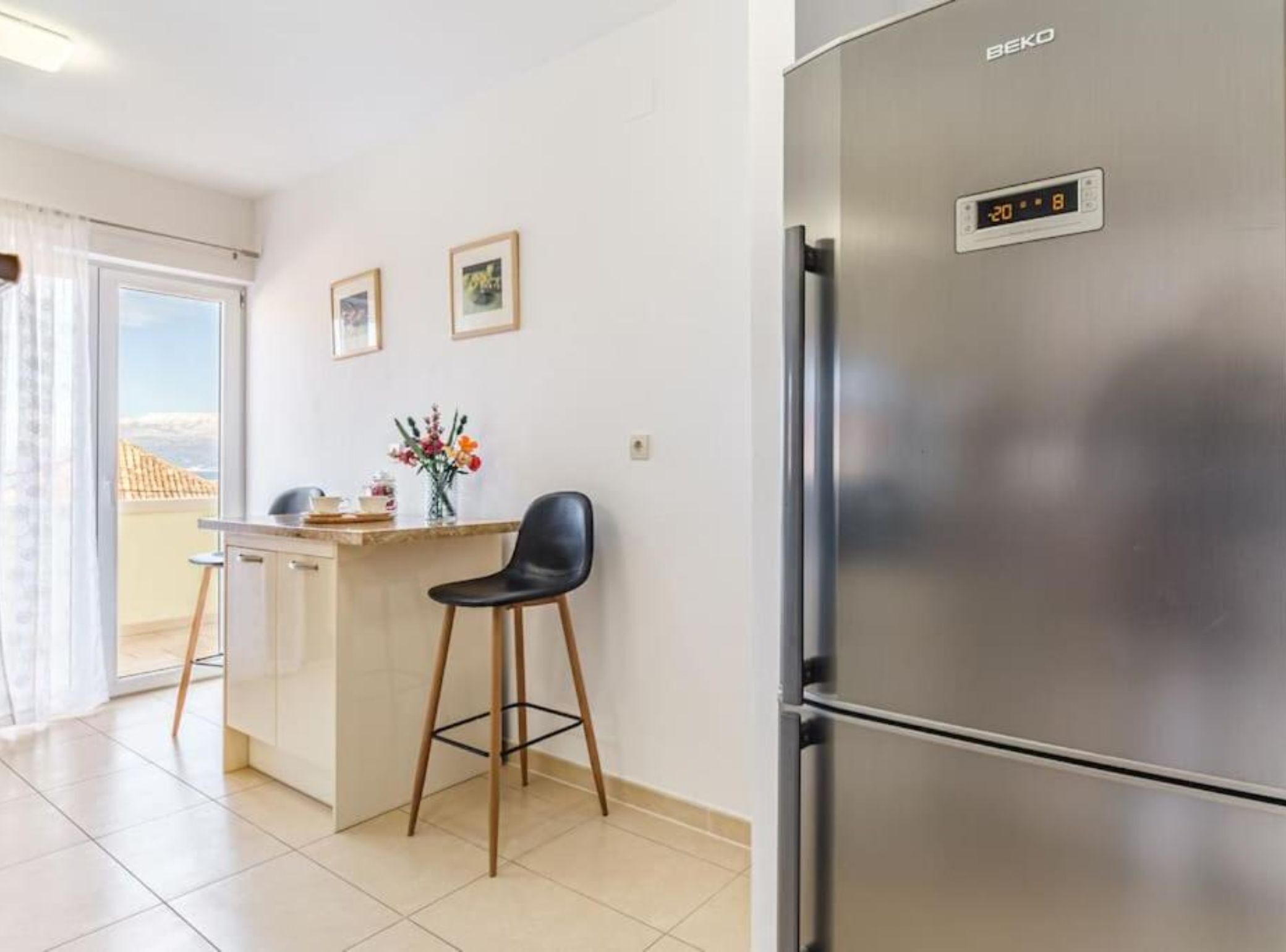 A modern kitchen with a stainless steel refrigerator, a small island with a chair, and a view through the glass door.