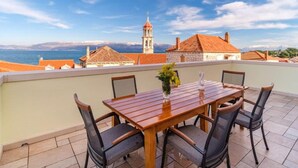 A terrace with a wooden table and chairs, a vase with flowers, and a view of a coastal town with a church tower. - Sutivan Sea Terrace Apartment (Sutivan)