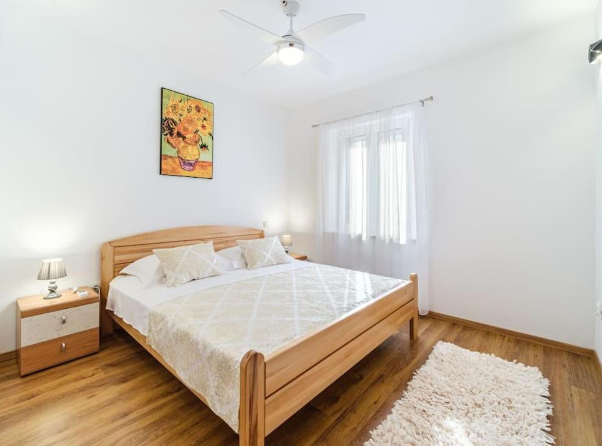 A bedroom with a wooden bed, a nightstand, a lamp, a ceiling fan, and a painting on the wall.