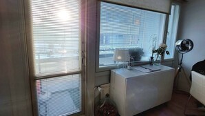 Et værelse med skrivebord, stol, radiator, et vindue med rullegardiner og en ventilator.