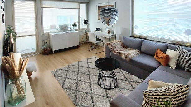 En moderne stue med en grå sofa, et sort sofabord og et tæppe med et mønster. Der er et skrivebord med en stol og en lampe i baggrunden.