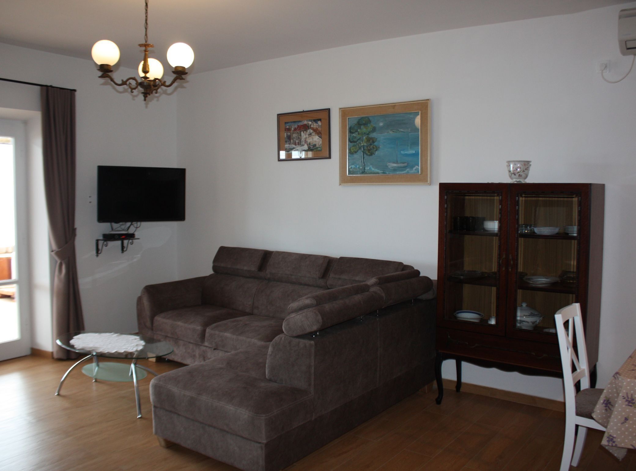 A living room with a sectional sofa, a glass coffee table, a television mounted on the wall, and a cabinet with dishes.