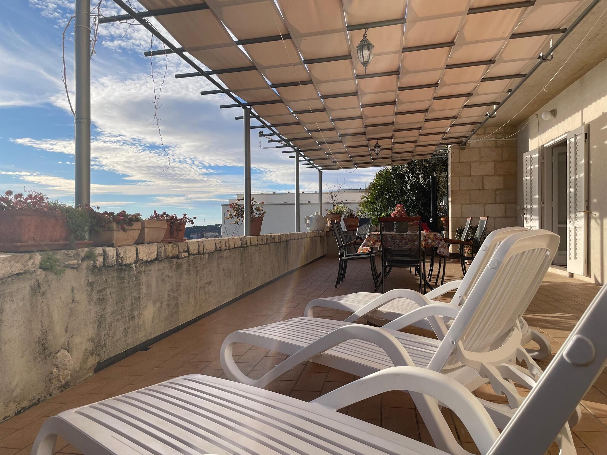 Un patio techado al aire libre con sillas y una mesa de plástico blanco, plantas en macetas y vista panorámica de la ciudad.