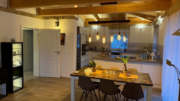 A modern kitchen with wooden beams, a dining area with a table and chairs, and a built-in bookshelf. - Bungalow "Villa Toscana" with Private Terrace, Garden, and Wi-Fi (Teltow)