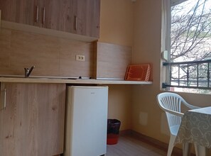 A compact kitchen with wooden cabinets, a white refrigerator, a small trash bin, and a window with a view of trees.