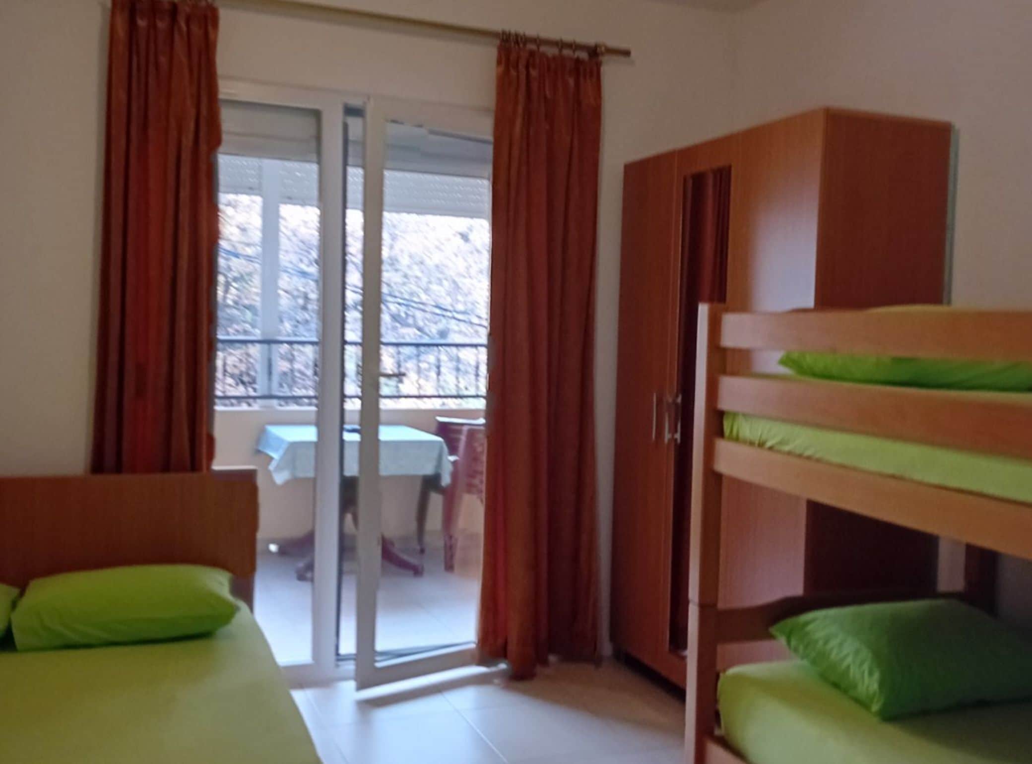 A dormitory room with bunk beds, a wooden wardrobe, and a sliding glass door leading to a balcony with a table and chairs.