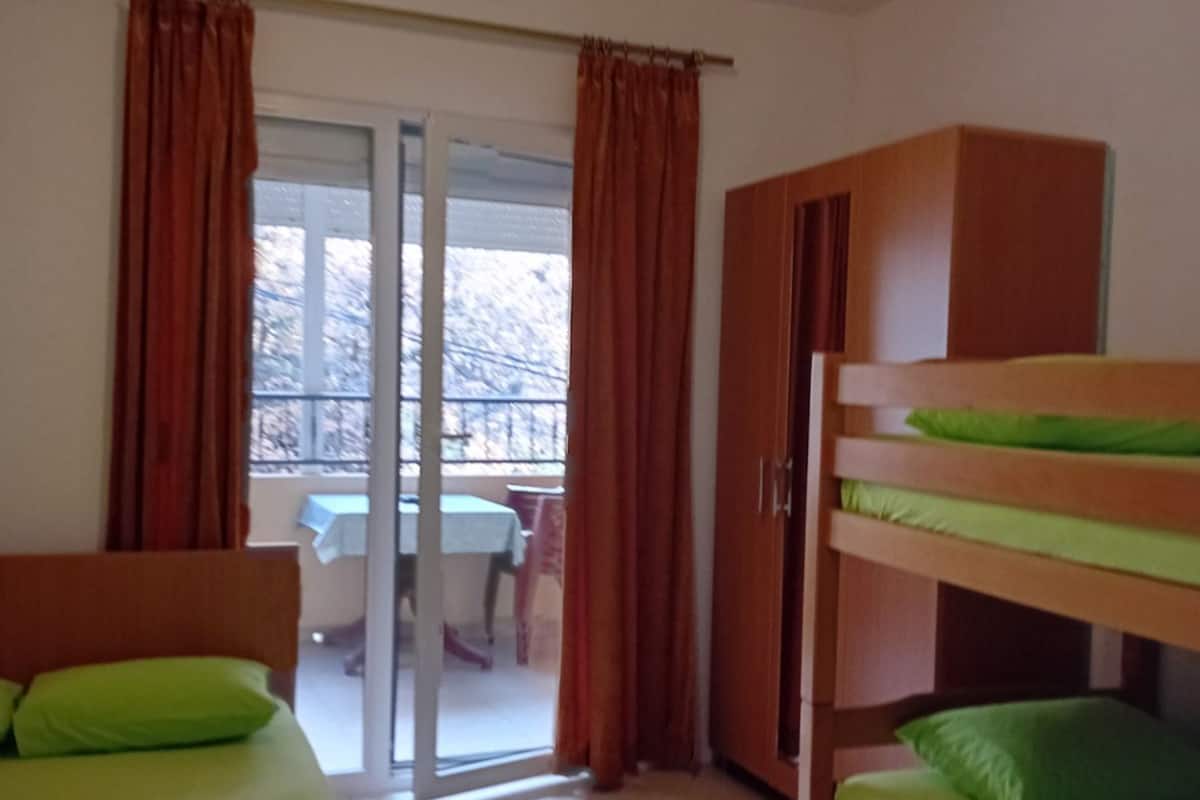 A dormitory room with bunk beds, a wooden wardrobe, and a sliding glass door leading to a balcony with a table and chairs.