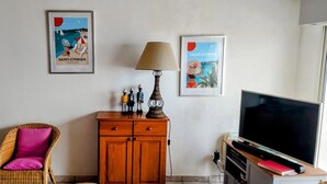 Habitación de hotel con una silla de mimbre, un armario de madera, una lámpara, un televisor y dos carteles enmarcados en la pared.