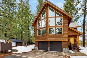 A two-story wooden cabin with large windows, a garage, and a staircase leading to the entrance. - Serene Creekside Chalet: Hot Tub, Games, BBQ (South Lake Tahoe)