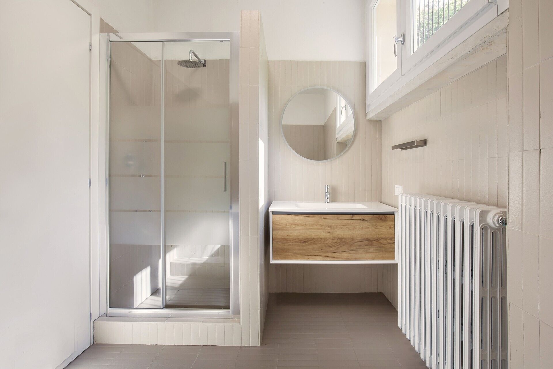 Un baño moderno con cabina de ducha de vidrio, tocador de madera, espejo redondo y radiador blanco.