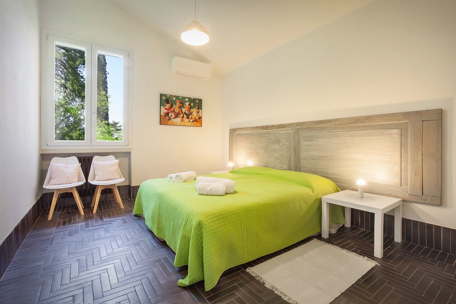 Une chambre avec une couvre-lit verte, deux tables de chevet, une tête de lit, une fenêtre donnant sur des arbres et un tableau au mur.