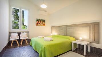 Une chambre avec une couvre-lit verte, deux tables de chevet, une tête de lit, une fenêtre donnant sur des arbres et un tableau au mur.
