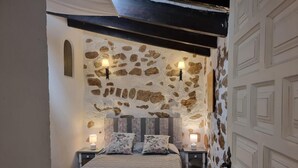 A bedroom with a stone wall, a bed with a quilted bedspread, two bedside tables with lamps, and a wooden ceiling beam. - Charming House, 2 Bedrooms, Terrace & Air Conditioning (L'Escala)