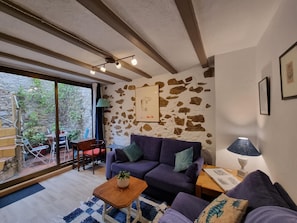 Ein gemütliches Wohnzimmer mit Steinwand, einem violetten Sofa, einem Holztisch und Blick ins Freie durch große Fenster.