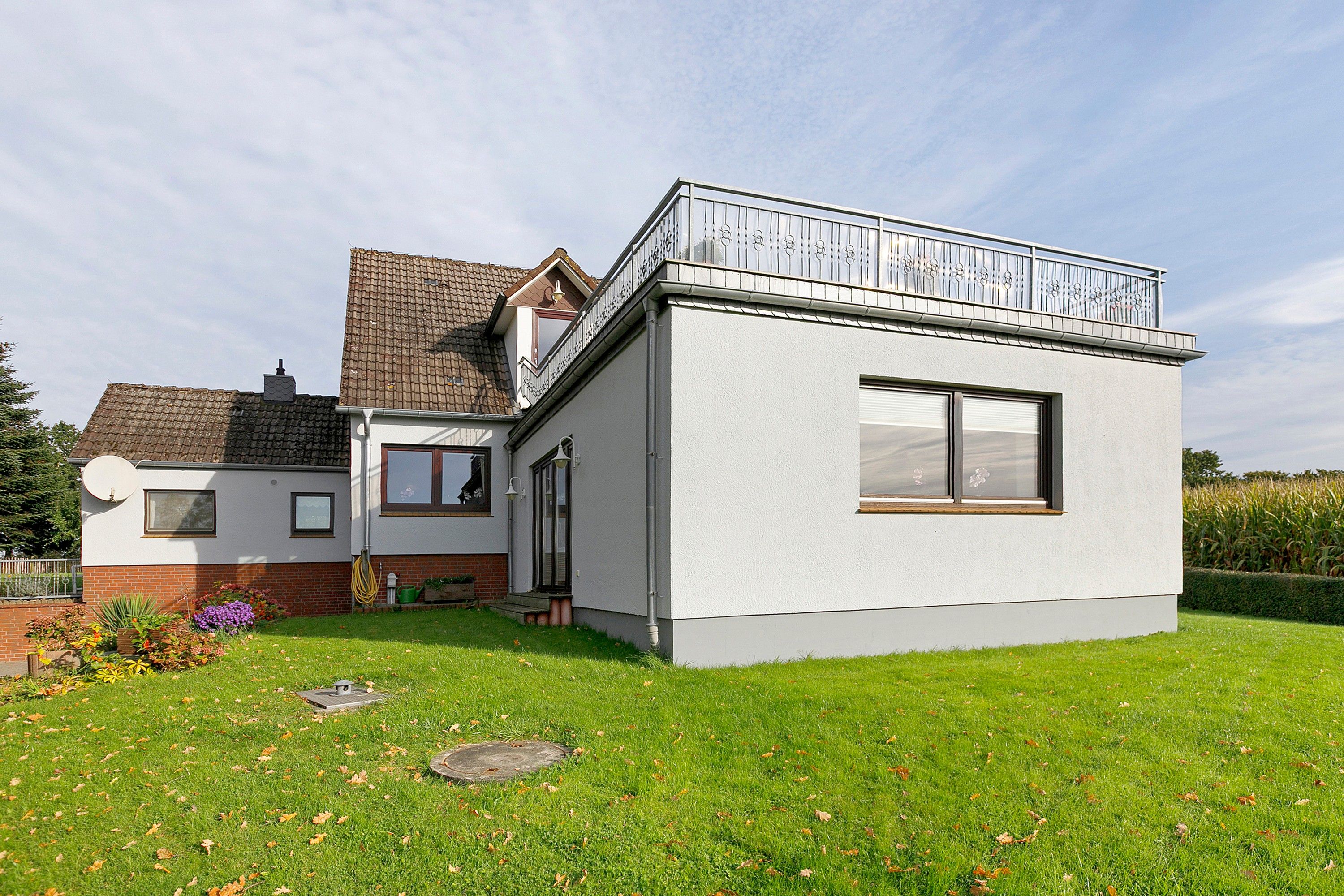 Une maison à deux étages avec un toit plat, un balcon doté d’une balustrade en métal et un petit jardin avec de la pelouse et des fleurs.
