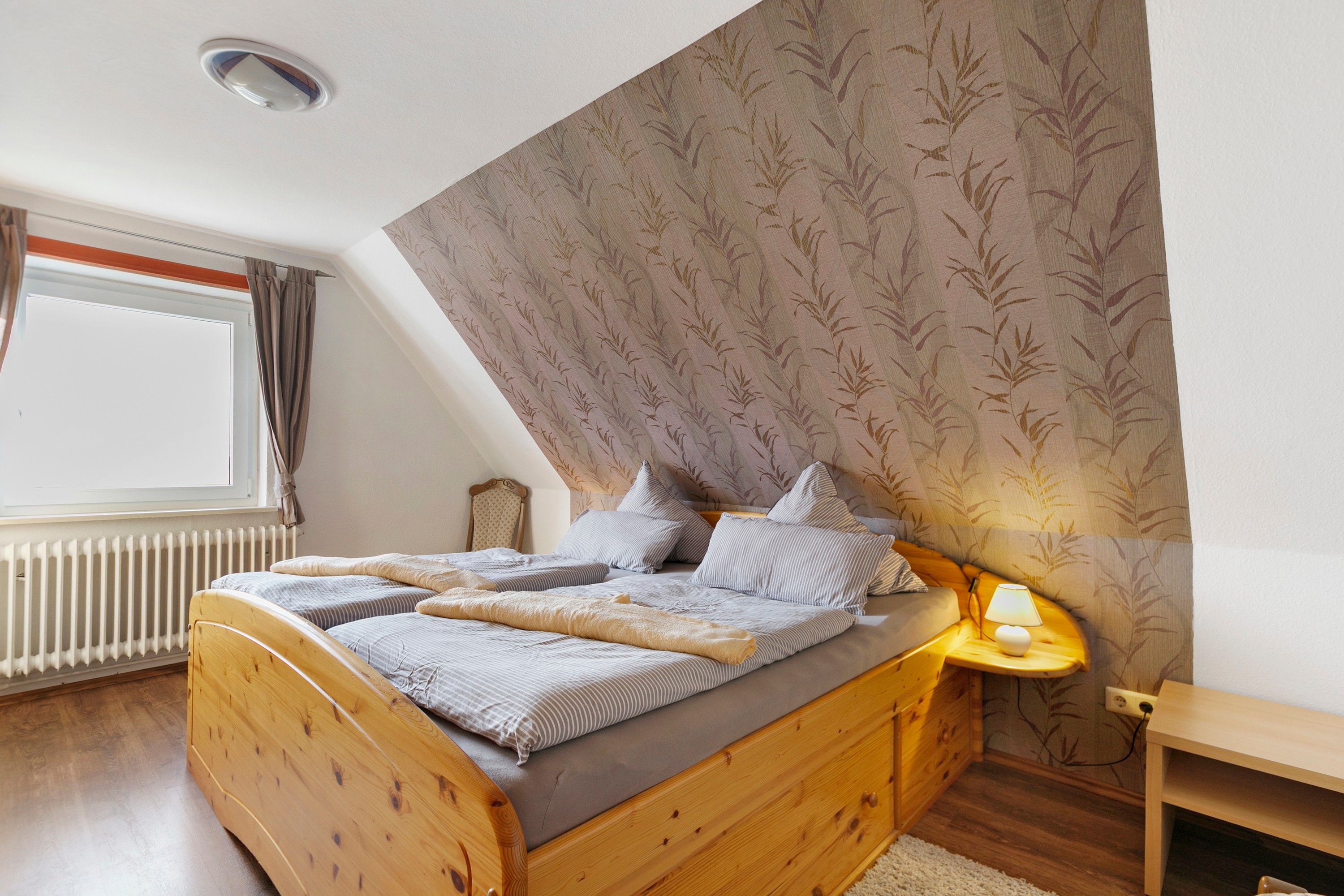 Ein Schlafzimmer mit einem hölzernen Bett, einem Nachttisch mit Lampe, einem Fenster mit Vorhängen und einer Heizung.