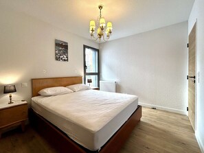 A bedroom with a wooden bed, a nightstand, a wall-mounted lamp, and a window with a view of the outdoors. - New 3-Room Apartment - 4 People - Terrace in Briançon (Briançon)