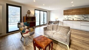 Ein modernes Wohnzimmer mit einer Couch, einem Sessel und einem Holztisch. Es gibt einen Essbereich mit Tisch und Stühlen sowie eine Küche mit weißen Schränken und einer Marmor-Spritzschutzwand.