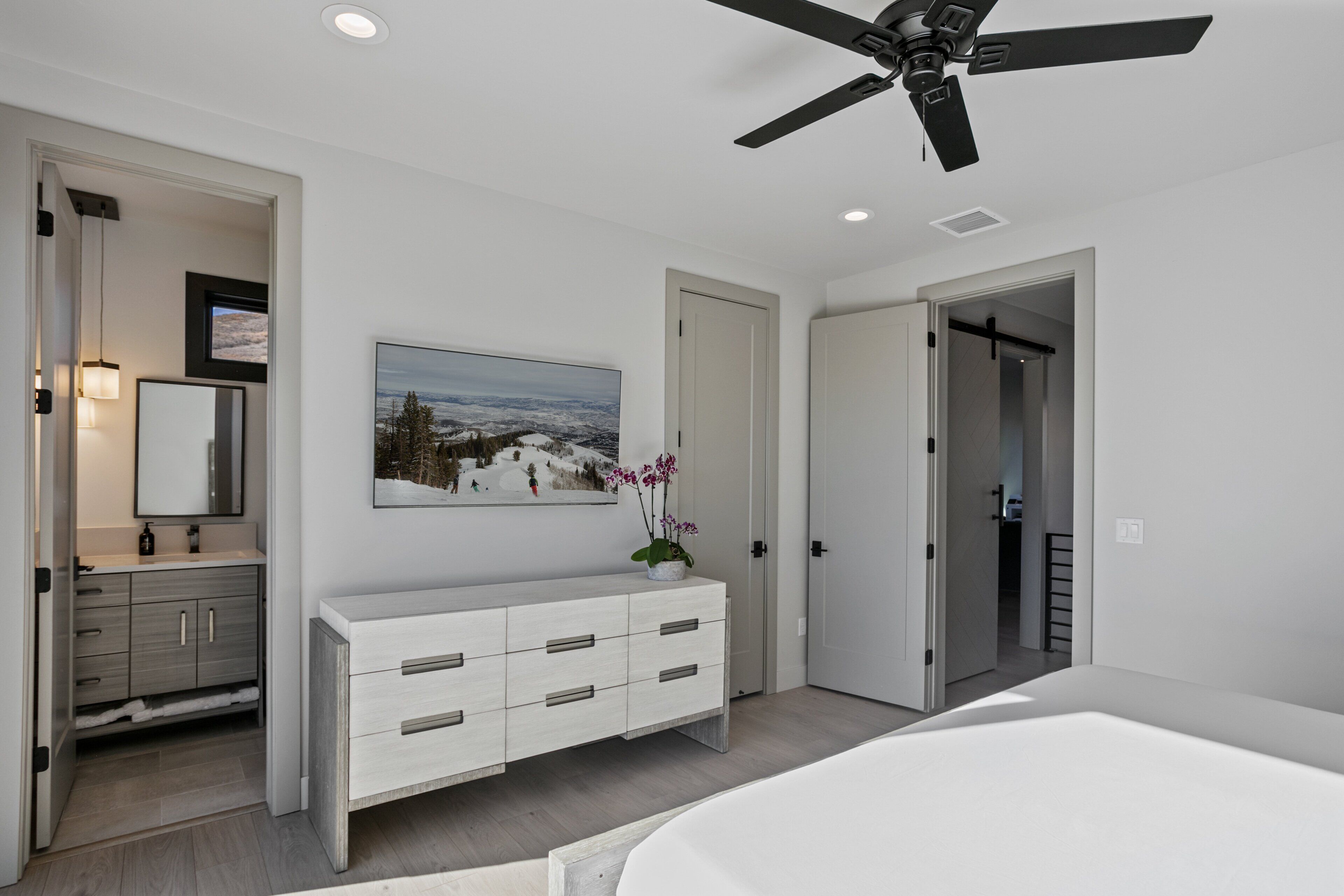 A bedroom with a ceiling fan, a large window, a white dresser, and a painting of a snowy landscape.