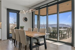 Ein moderner Essbereich mit Holztisch und Stühlen, großen Fenstern mit Blick ins Freie und einer Wanduhr.