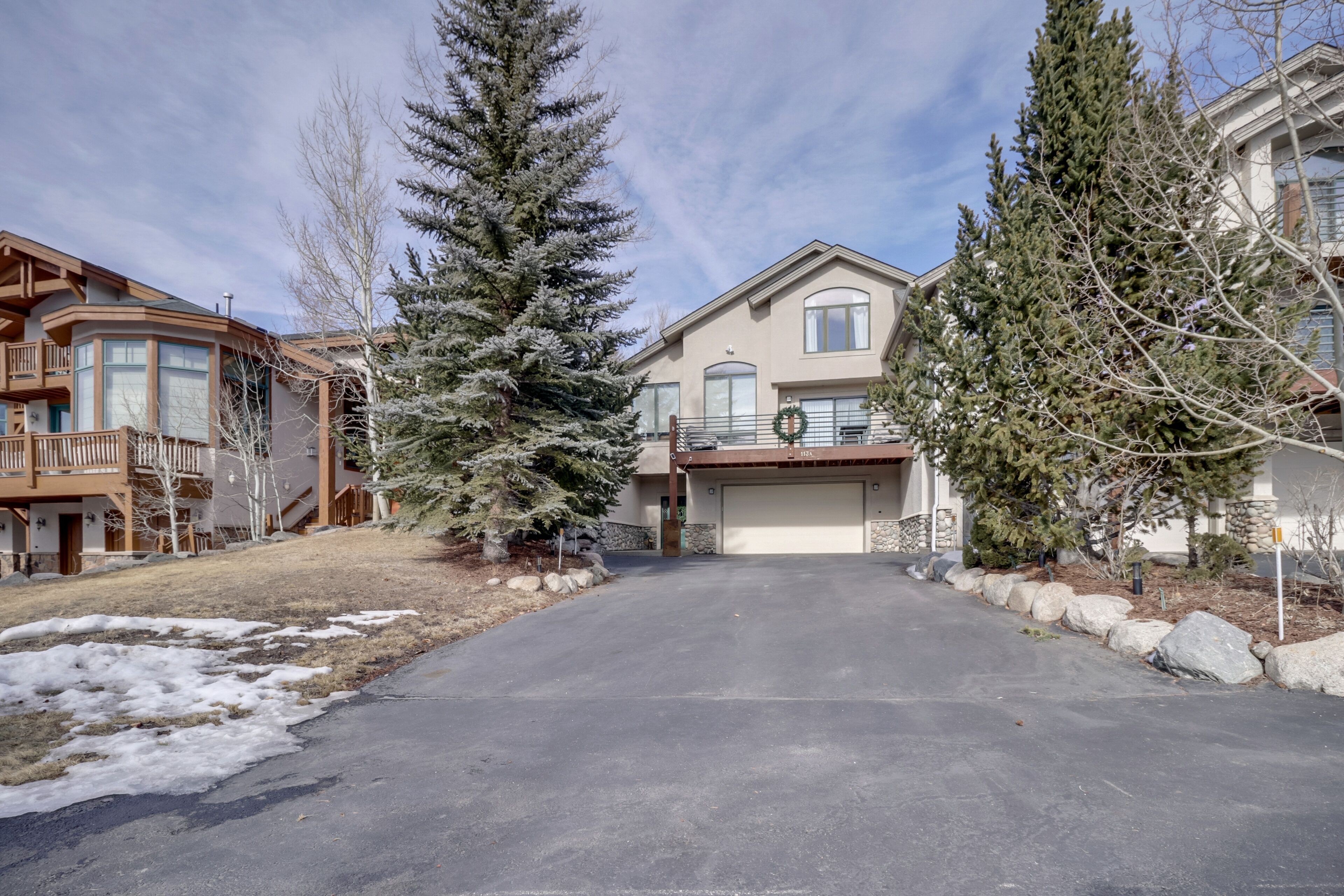 A residential driveway leading to a two-story house with a garage, surrounded by snow-covered ground and evergreen trees.