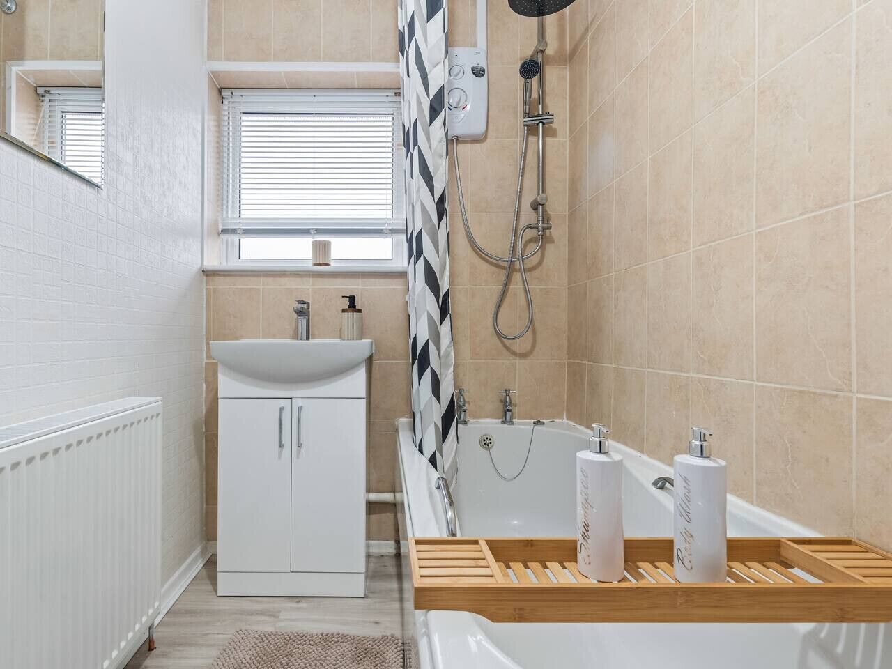 A bathroom with a white sink, a bathtub, and a shower. There is a window with blinds, a radiator, and a wooden tray with toiletries.