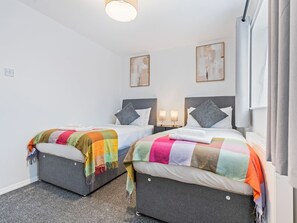 Ein Hotelzimmer mit zwei Betten, jeweils mit bunter Decke und Kissen, einem kleinen Nachttisch mit Lampe und einem an der Wand montierten Fernseher.