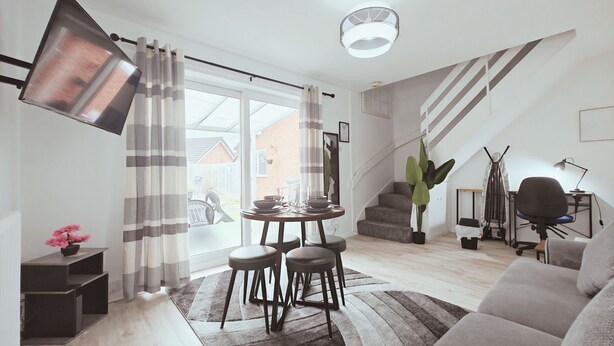 A modern living room with a sofa, a dining table with stools, a staircase, and a large window. - Telford Centre House with Parking, Garden, Wi-Fi (Telford)