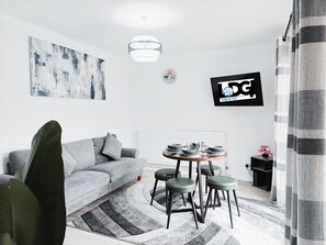 Ein modernes Wohnzimmer mit einem grauen Sofa, einem runden Esstisch mit Hockern, einem hängenden abstrakten Gemälde, einer Uhr und einem Fernseher, der ein Logo zeigt.