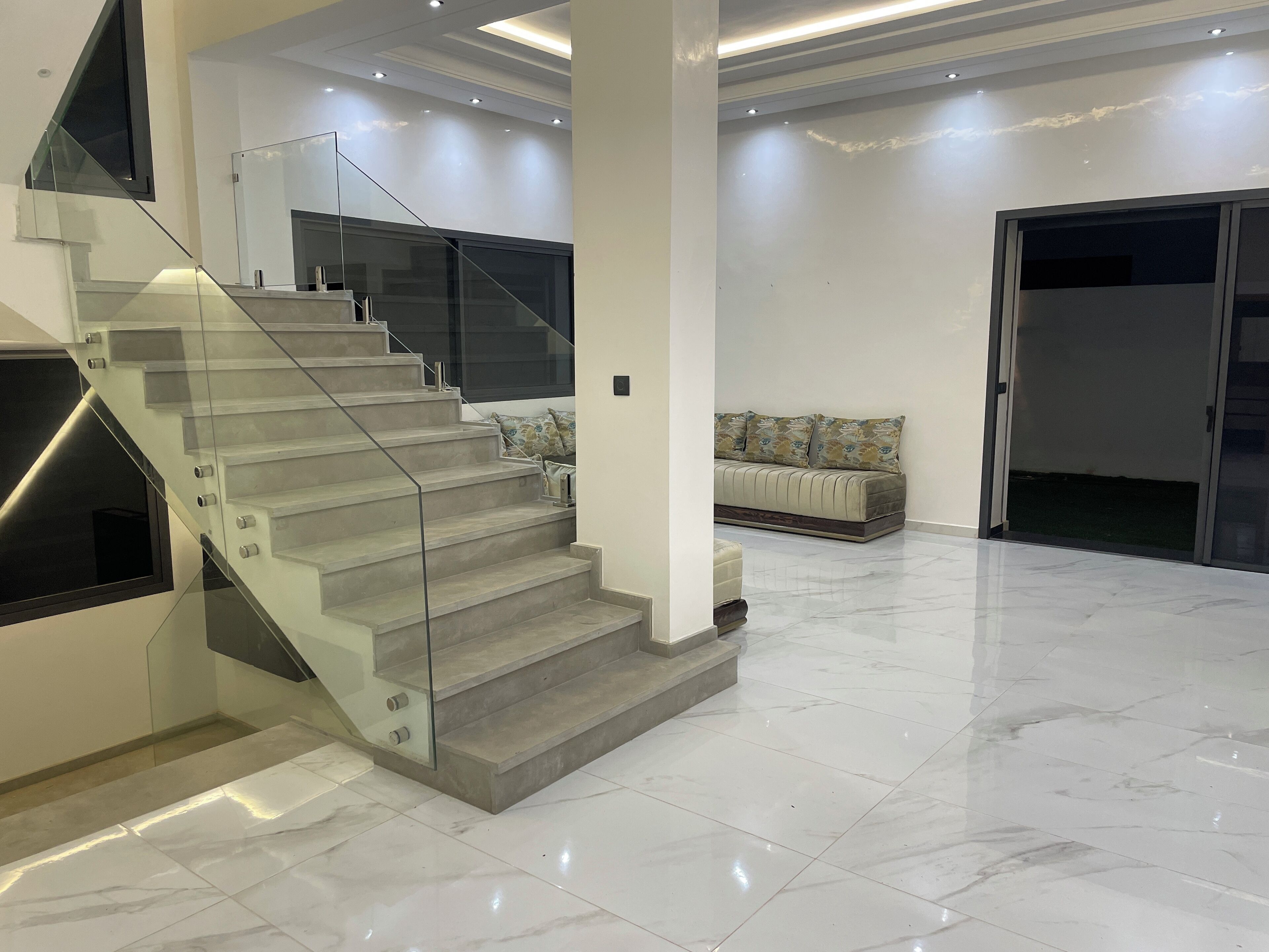 Een modern interieur met een glazen en metalen trap, een bank met patroon kussens en een strakke, betegelde vloer.
