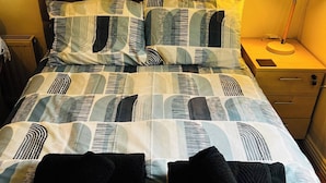 Sängyssä on kuviollinen päiväpeite ja kaksi mustaa tyynyä, yöpöydällä on lamppu ja sängyn vieressä on puinen yöpöytä, jossa on yksi laatikko.