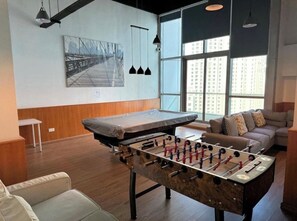 A room with a foosball table, a sofa, and a large window with a view of buildings. - Pool & hot tub, amazing Marina location steps to metro & JBR beach - 1BR (Dubai)