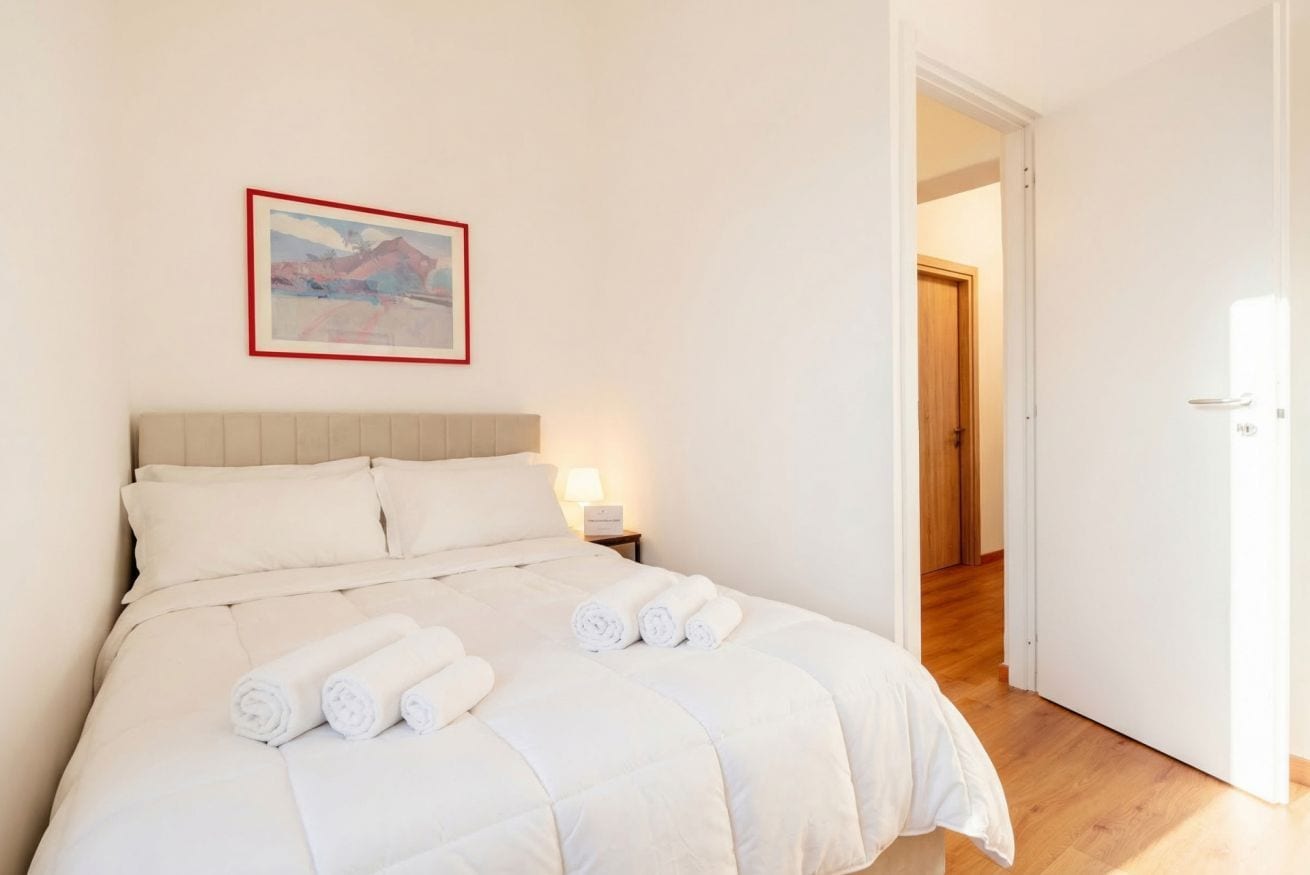 A neatly made bed with rolled towels, a framed picture on the wall, and a wooden door in the background.