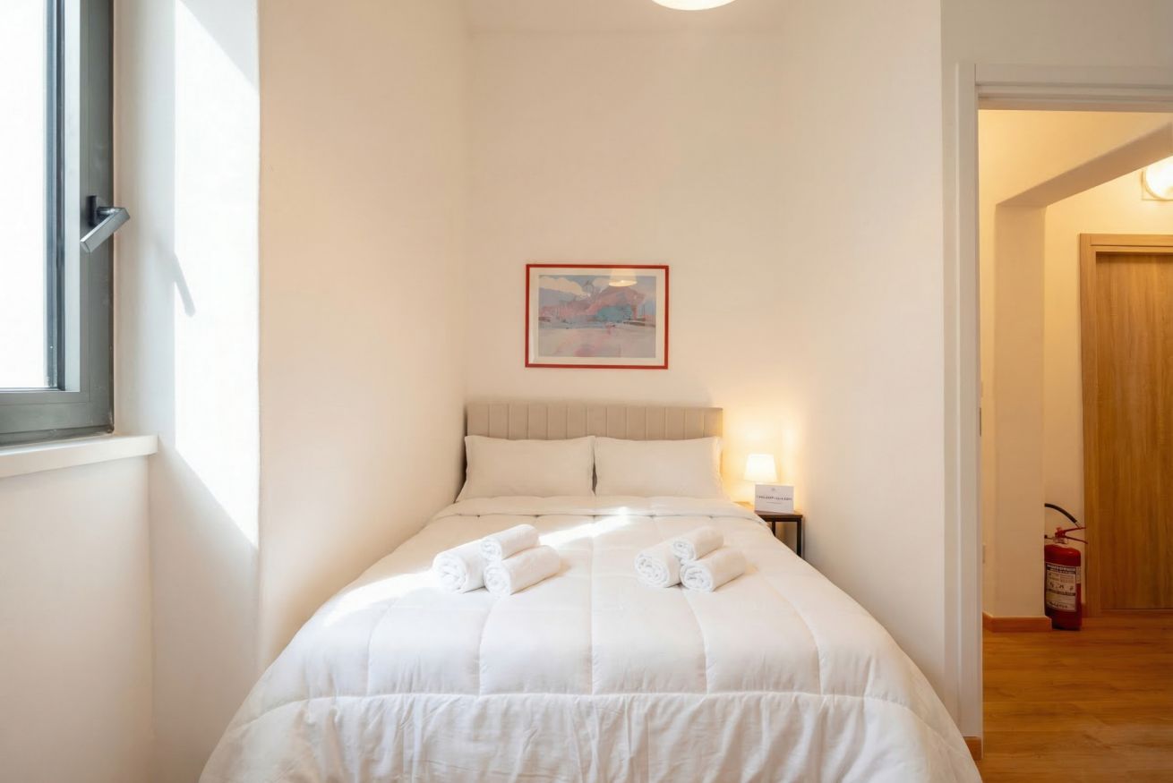 Uma cama bem arrumada com toalhas dobradas, um quadro na parede e um extintor de incêndio no corredor.