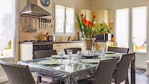 Un'area pranzo con tavolo in vetro, sedie in vimini, una cucina con utensili appesi sopra e un orologio a parete.