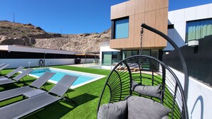 Area piscina moderna all'aperto con poltrone prendisole, una sedia a dondolo e un edificio con ampie vetrate.