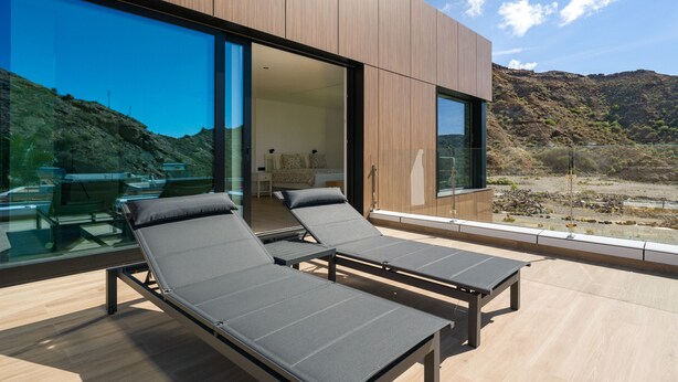 Un'area lounge esterna moderna con due lettini prendisole e una stanza con pareti in vetro visibile attraverso le porte scorrevoli.