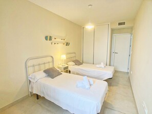A hotel room with two single beds, a bedside table with a lamp, a wall-mounted shelf with decorative items, and a door leading to another room. - Modern Ground Floor Apartment in Islas Menores with Community Pool and Garden (Islas Menores)