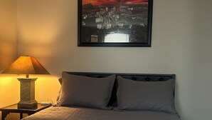 Un dormitorio con cama, mesita de noche, lámpara y un cuadro con una vista urbana enmarcada en la pared.