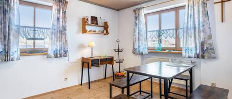 Una habitación con suelo de madera, una mesa con bancos, una lámpara, una estantería con adornos y dos ventanas con cortinas.