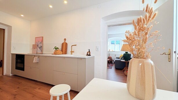 Una cocina moderna con una isla central, una zona de comedor con una mesa blanca y un jarrón de flores secas, y vistas al salón.