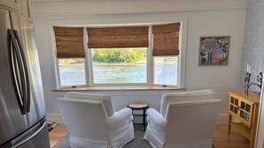 A kitchen with a refrigerator, two white chairs, a small round table, and a window with blinds overlooking a body of water. - Stunning 3-bedroom Weskeag Village River House in South Thomaston (South Thomaston)