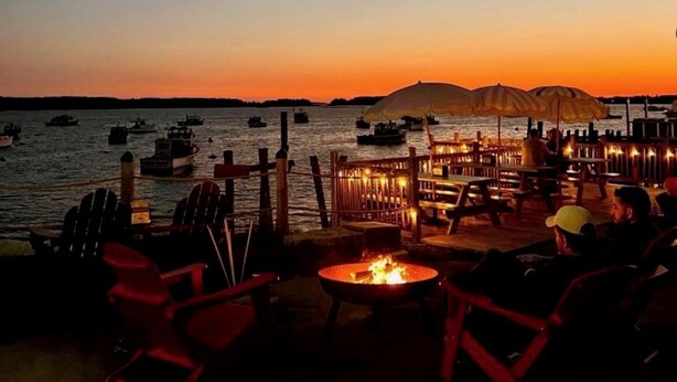 An outdoor seating area with red chairs and a fire pit, overlooking a body of water with boats and a dock in the background. - Stunning 3-bedroom Weskeag Village River House in South Thomaston (South Thomaston)
