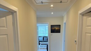 A hallway with white walls, wooden flooring, and white doors. There is a radiator on the left, a picture on the wall, and a window with blinds in the background.