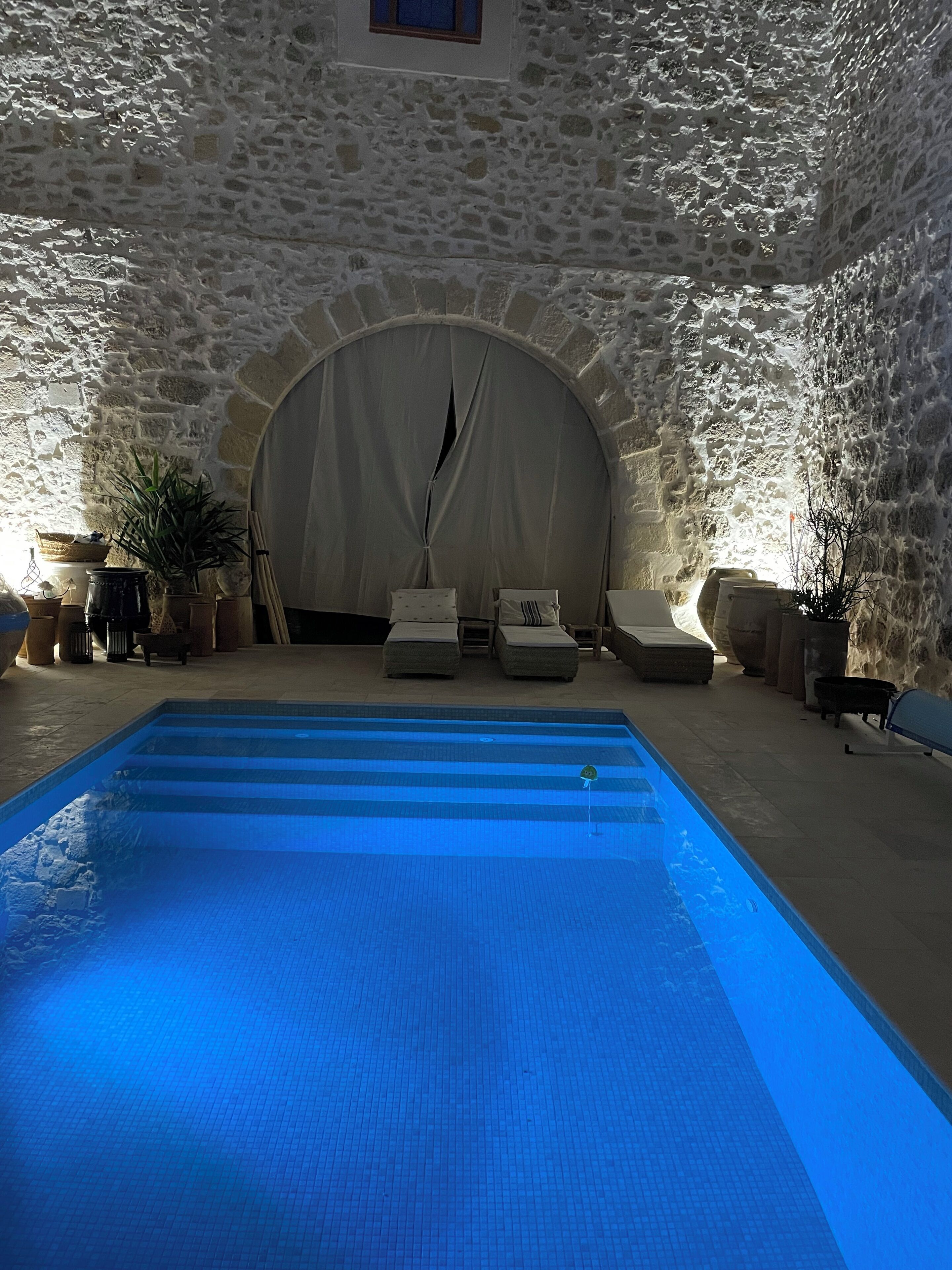 Une piscine intérieure aux murs de pierre, avec une eau d’un bleu limpide, entourée de chaises longues et de plantes en pot.