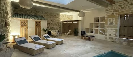 Un espace aménagé au bord de la piscine, avec des chaises longues, un mur en pierre et un plafond avec des poutres apparentes.