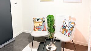 Uma sala de estar moderna com dois poltronas brancas, uma mesa de centro preta e uma luminária pendente. Há duas pinturas abstratas na parede e um vaso com planta entre as poltronas.