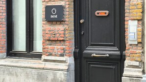Une porte noire avec une poignée en laiton, une enseigne noire sur laquelle est écrit le mot « nid », et un mur de briques.