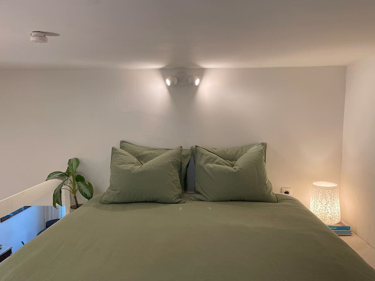 Un letto con due cuscini, una lampada da comodino e una pianta in vaso in una stanza con pareti bianche e un lampadario.