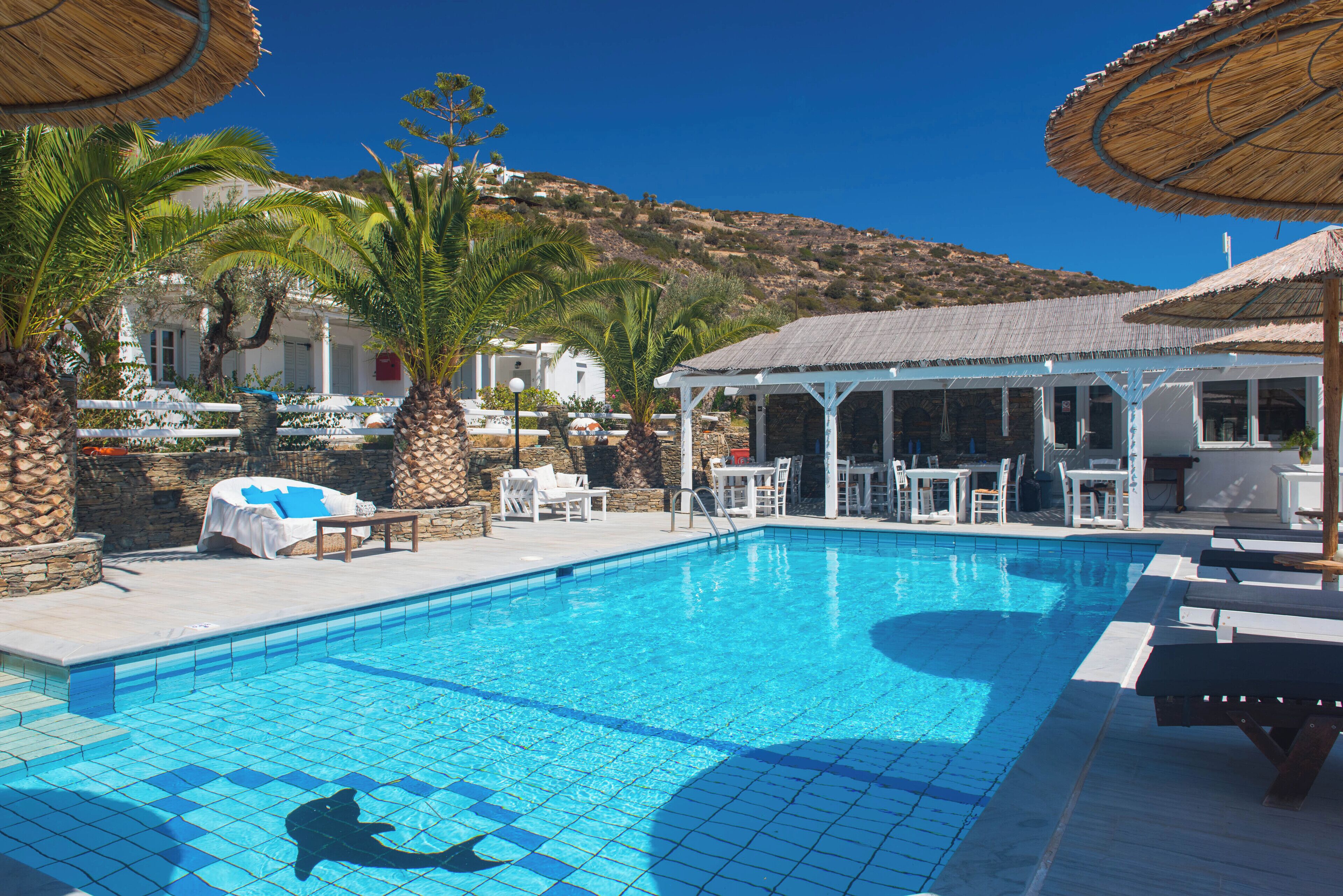 A swimming pool with lounge chairs and umbrellas, surrounded by palm trees and a building with a white facade.